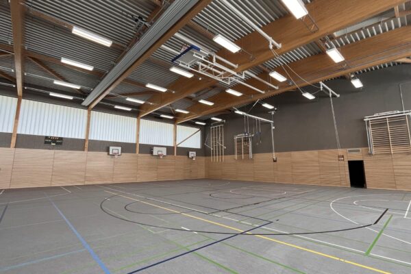 Bild zeigt die neue Dreifeld-Sporthalle am Niekampsweg, die auf einem Holzbau basiert. Die Prallwände rundherum sind aus Holz, auch das Dach besteht aus einer offen sichtbaren Holzkonstruktion.
