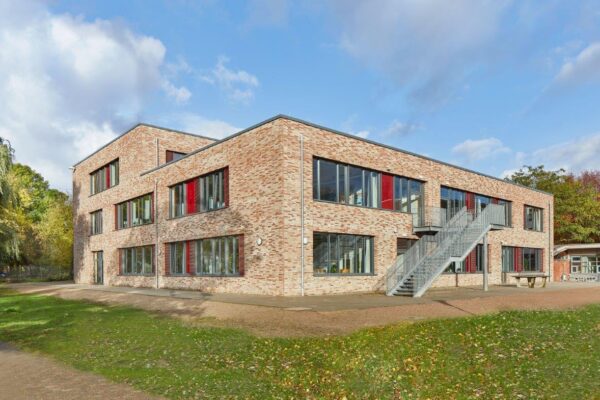 Außenansicht der Stadtteilschule Wilhelmsburg. Gebäude aus hellen Ziegeln mit teils farbigen Fensterlementen. Davor eine Grünfläche, im Hintergrund Bäume.