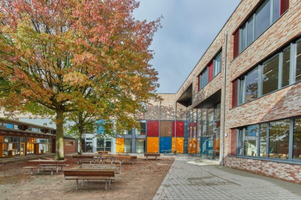Außenansicht der Stadtteilschule Wilhelmsburg. Gebäude aus hellen Ziegeln mit teils farbigen Fensterlementen. Davor ein Innenhof mit Bäumen.